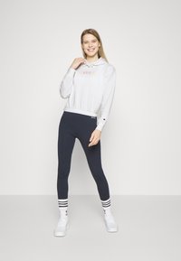 Biela mikina s nápisom "CHAMPION", elastický pás, skombinovaná s tmavými námorníckymi legínami a bielymi teniskami so pruhovanými ponožkami. Minimalistické pozadie.