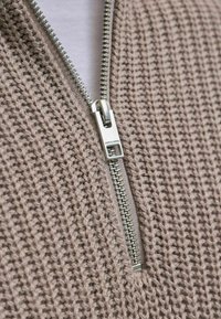 Prenda de punto beige con cremallera, adornada con una cremallera metálica plateada. Presenta un tejido con textura y patrones verticales, y un interior forrado en blanco.