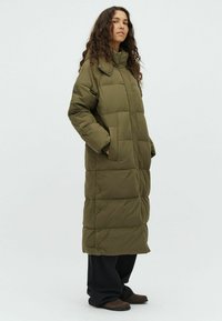 Woman standing wearing a long olive green puffer coat with hands in pockets, black pants, and brown shoes against a plain light background.