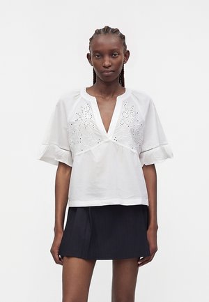 Γυναίκα με πλεξούδες φορώντας λευκό κεντητό πουκάμισο με φαρδιά μανίκια και βαθύ V-λαιμό, σε συνδυασμό με κοντή μαύρη φούστα σε λευκό φόντο.