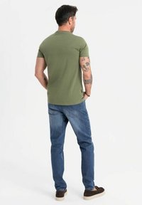 Camiseta de manga corta en verde oliva, diseño ajustado, combinada con jeans azules. La textura muestra un acabado liso. Unos zapatos marrones completan el conjunto.