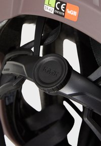 Pokret pokrętła regulacji kasku w czarnym plastiku, o gładkiej fakturze. Przypięte paski w czerni, otoczone brązową powłoką kasku.