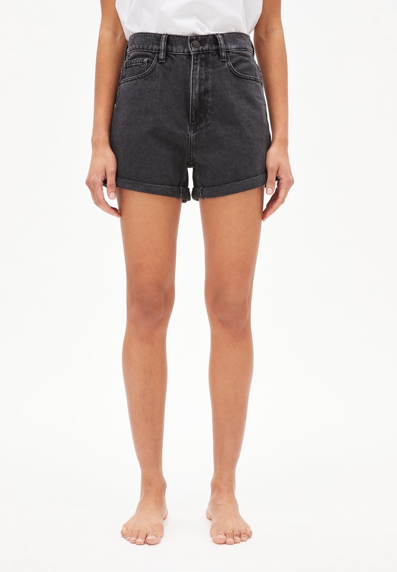 Person som bär upprullade svarta jeansshorts och en vit topp, står barfota mot en enkel vit bakgrund.