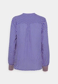Blusa de manga larga en azul con grandes lunares blancos. Presenta un escote redondo, una tapeta de botones y puños a rayas con acentos en rojo y blanco.