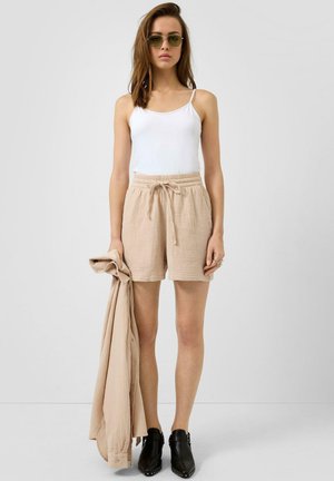 Kobieta w zielonych okularach przeciwsłonecznych, białej bluzce na ramiączkach, beżowych szortach ze sznurkiem, czarnych butach, trzymająca beżową, dobraną koszulę z długim rękawem na białym tle.