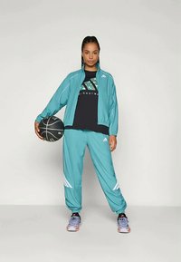 Completo sportivo teal con dettagli bianchi, composto da una giacca con zip e pantaloni larghi. Maglietta nera con grafica. Tiene in mano un pallone da basket.