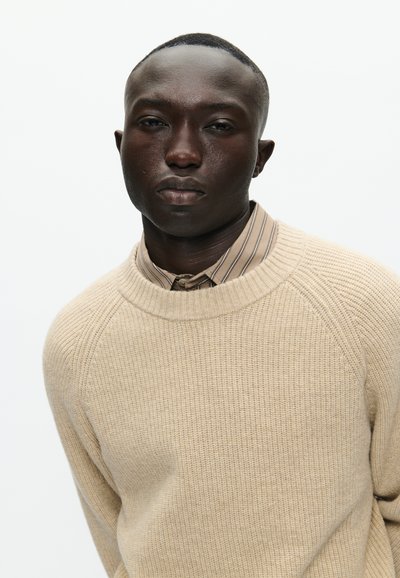 Bežani pleteni pulover s rebrastom teksturom i širokim okruglim izrezom, u kombinaciji s uskim prugastim majicom u neutralnim tonovima.