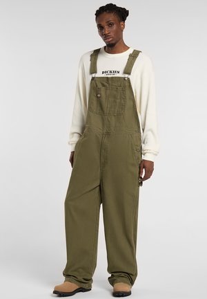 Olivgrüne Baumwoll-Latzhose mit verstellbaren Trägern, einer Fronttasche und weitem Bein, kombiniert mit einem weißen Langarmshirt.