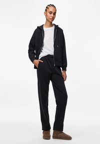 Juodas zip-up džemperis su priekinėmis kišenėmis, derinamas su juodomis sportinėmis kelnėmis. Modelis po juo dėvi baltą marškinėlį. Rudos aulinės botinės užbaigia įvaizdį.