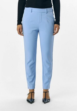 Woman wearing light blue fitted trousers paired with black long-sleeve top and black pointed-toe T-strap heels, standing straight.