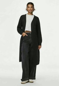 Μακριά μαύρη κάρντιγαν από μαλακό ύφασμα, με ανοιχτό μπροστινό σχέδιο, συνδυασμένη με άσπρη μπλούζα και ψηλόμεσα μαύρα τζιν παντελόνια.