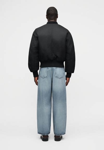 Άνδρας με την πλάτη, φοράει μαύρο bomber μπουφάν, ανοιχτό μπλε τζιν με φαρδιά πόδια και μαύρα παπούτσια, σε απλό λευκό φόντο.