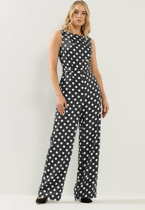 Kvinde iført sort ærmeløs jumpsuit med hvide polkaprikker, bælte i taljen og bukser med brede ben, stående foran en ensfarvet lys baggrund.