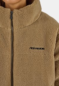 Giacca in pile marrone con colletto alto, dotata di zip centrale e logo ricamato nero sul petto. Morbida al tatto.