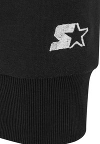 Puño de tela negra con la letra "S" bordada en blanco, parcialmente cubierta por una estrella, textura de punto y borde acanalado visibles.