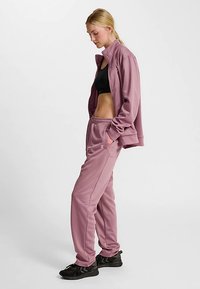 Paarse sportieve trainingspak met een jas met rits en broek met elastische taille. Materiaal lijkt glad met een subtiele glans. Zwarte sportbeha eronder.