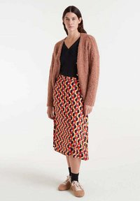 Cárdigan marrón de felpa sobre una camiseta negra, combinado con una falda midi de patrón rojo, amarillo y negro y zapatillas marrón claro con cordones blancos.