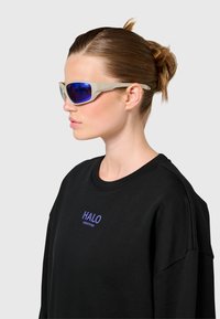 Kobieta z jasnobrązowymi włosami upiętymi w kok, nosząca beżowe okulary przeciwsłoneczne z niebieskimi soczewkami i czarną bluzę z napisem "HALO" na szarym tle.