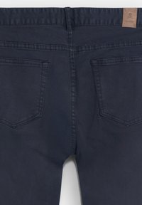 Vaqueros de mezclilla en azul oscuro con una textura suave, dos bolsillos traseros y una etiqueta de cuero en la cinturilla. Diseño simple con un detalle mínimo.