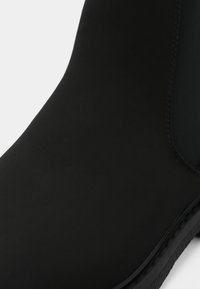 Mustat nilkkurit, jotka on valmistettu sileästä materiaalista. Niissä on pyöreä nenä, elastiset sivupaneelit ja teksturoitu kontrastiväri pohja.