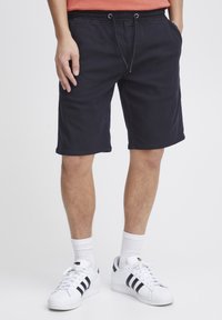 Svarta bomullsshorts med elastisk midja, snörning, sidofickor och en avslappnad passform. Bärs med vita sneakers och strumpor.