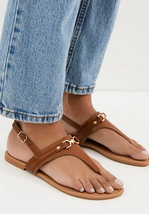 Pieds chaussés de sandales plates en cuir marron avec des détails de boucle dorée, accompagnées d'un jean denim bleu clair.