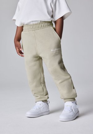 Niño que lleva puestos pantalones deportivos beige de AllSaints, zapatillas blancas y camiseta blanca, de pie frente a un fondo gris liso.