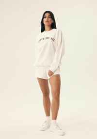 Biała bluza z napisem „COSTA DEL SOL” w bordowym kolorze, połączona z dopasowanymi białymi szortami. Model nosi białe sneakersy i skarpetki do kostek.