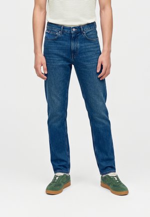 Personne portant un jean bleu droit, des baskets vertes en daim avec des semelles en gomme, et une chemise blanche rayée, debout devant un fond uni.