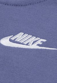 Fialová bavlnená tkanina s bielou vyšitou logom Nike, s zakriveným dizajnom a textúrovanou väzbou. Detailný pohľad zvýrazňuje šitie.