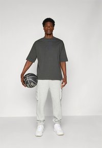 Mørkegrå T-skjorte med korte ermer har en subtil logo; kombinert med lysegrå cargo-sweatpants og hvite sportssko. Holder en svart basketball.