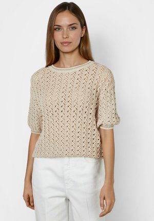 Young woman wearing beige open-knit short-sleeve sweater and white pants, standing against a plain light background.