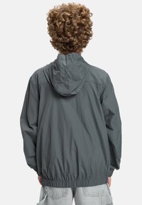 Grå vindtät jacka med huva, elastisk midja och ärmslut. Har ett lätt, skrynkligt tyg med en mjuk textur.