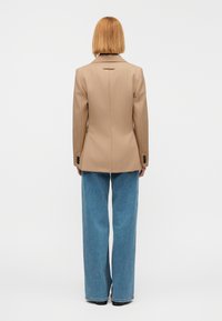 Μπεζ σακάκι κατά παραγγελία με εφαρμοστή δομή, μία σχισμή στο πίσω μέρος και μαύρα κουμπιά ως διακοσμητικά, συνδυασμένο με φαρδιά ανοιχτό μπλε τζιν παντελόνια.