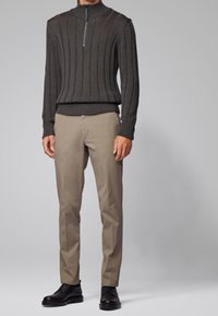 Pull gris foncé côtelé avec un col montant et une fermeture éclair, associé à un pantalon beige et des chaussures en cuir noir, créant un look en couches.