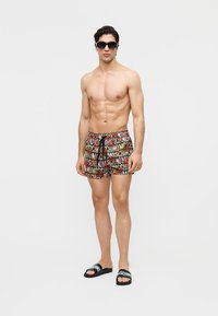 Homme torse nu portant un short multicolore de la marque Moschino, des sandales noires Moschino et des lunettes de soleil noires, debout devant un fond blanc.