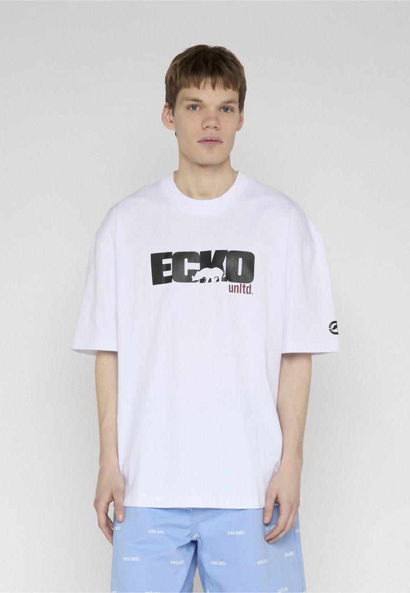 Jeune homme portant un T-shirt blanc oversize de la marque Ecko Unltd. et un short bleu clair avec des logos répétés d'Ecko Unltd., debout devant un fond uni.