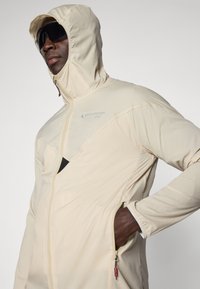 Beige lightweight jacket with a hood, zipper closure, and mesh accents. Features a logo on the chest and a pocket on the side.