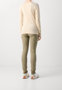 Beige shirt met lange mouwen, olijfgroene slim-fit broek en witte sportschoenen met oranje accenten, gezien van achteren tegen een lichte achtergrond.
