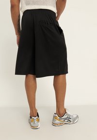 Shorts de sport noirs avec une taille élastique et des poches latérales, associés à des chaussures de course argentées et dorées, vus de dos.
