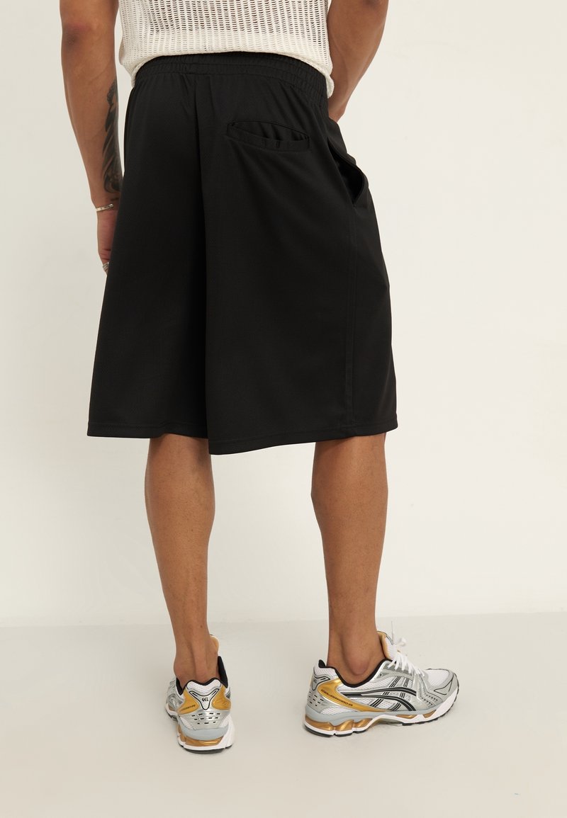 Shorts de sport noirs avec une taille élastique et des poches latérales, associés à des chaussures de course argentées et dorées, vus de dos.