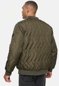 Chaqueta bomber acolchada de color verde oliva con cuello y puños acanalados, que presenta un patrón de diamantes y un parche de logo negro en el hombro.