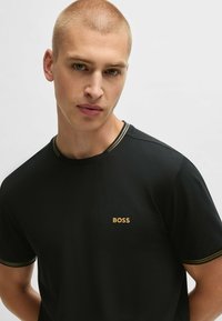 Juodas marškinėlis su apykakle, ant krūtinės turi auksinį "BOSS" logotipą ir dviejų tonų juostelės detales ant apykaklės. Lygi tekstūra.