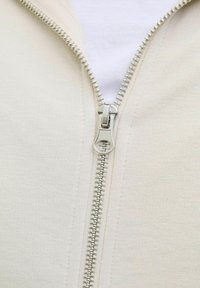 Bež hoodie na zip s glatkom teksturom, s metalnim munjom i bijelim unutarnjim tamplom, naglašen suptilnim končanim detaljima.
