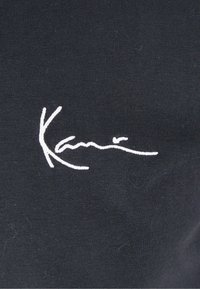 Svart stoff med en glatt tekstur som har en hvit brodert signatur. Designet er enkelt, og fremhever logodetaljen.