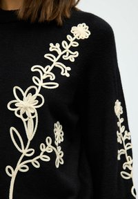 Pull noir avec un délicat broderie florale couleur crème sur le devant et les manches, d'une texture douce et d'une coupe décontractée.