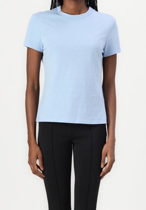 Person wearing a light blue short-sleeve crew neck t-shirt and black fitted pants, standing against a plain white background.