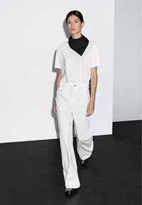 Bílá trička a široké bílé kalhoty, doplněné černým šátkem. Oblečení má uvolněný střih a jemnou texturu látky.