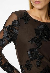 Juodas permatomas viršus su sudėtingu gėlių siuvinėjimu, kuriame derinamos skirtingos tekstūros ir formos, ant tamsiai rudos medžiagos fono.