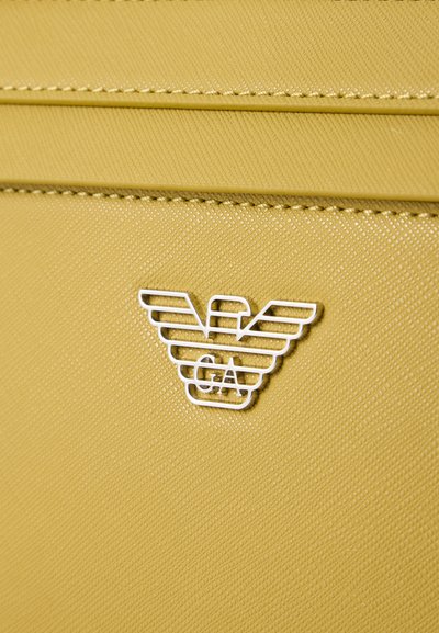 Close-up of textured yellow leather with a silver eagle-shaped logo featuring the letters V and GA on a bag or accessory.